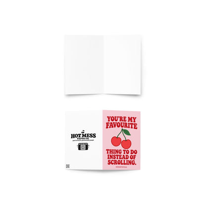 Funny romantic greeting card featuring two bright red cherries connected by green stems with a single leaf on a soft pink background. Bold red retro lettering reads “You're My Favourite Thing to Do Instead of Scrolling.” Sweet love card celebrating less screen time, more quality time, and real-life connection by Hot Mess Paper Co.