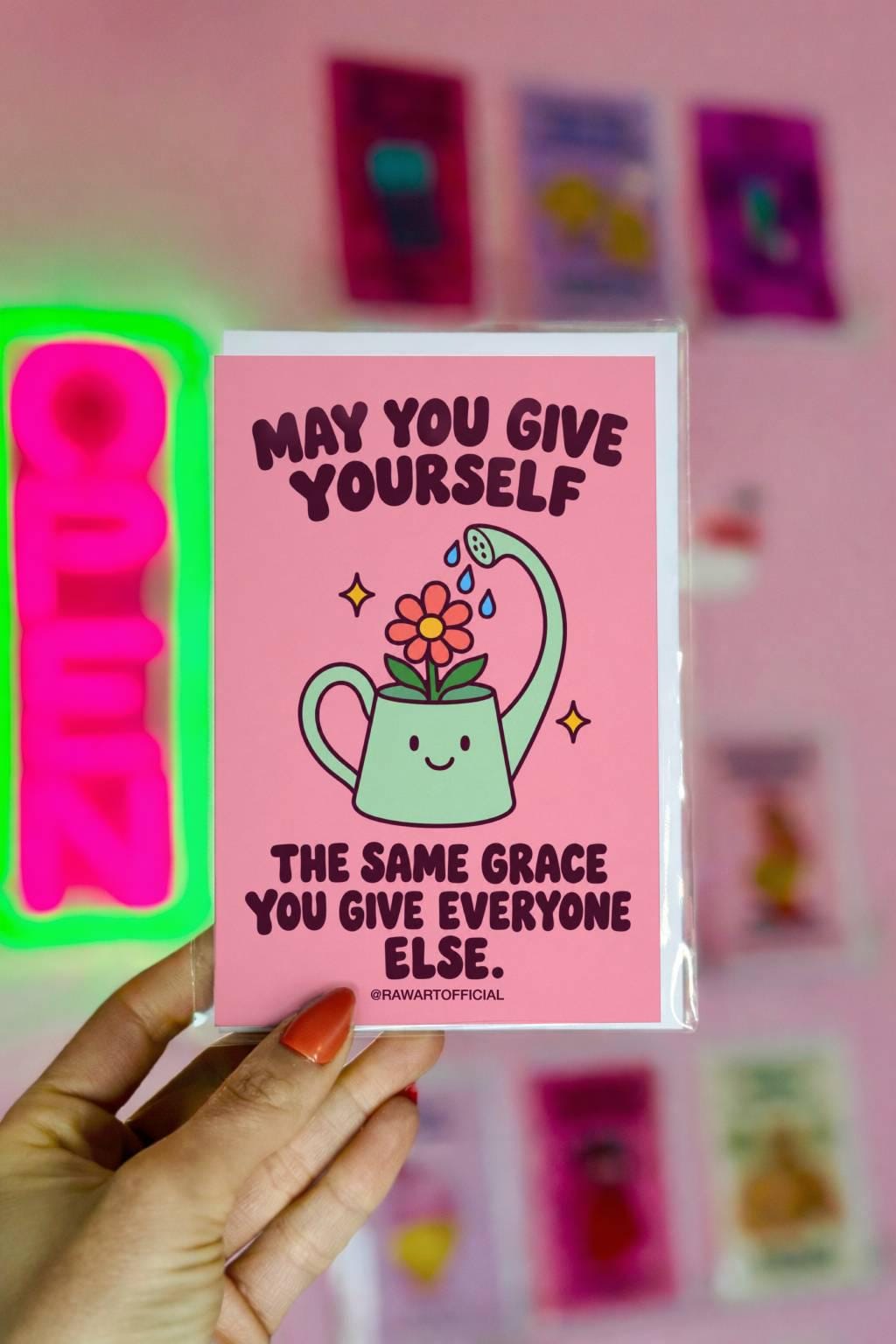 Heartfelt encouragement greeting card featuring a smiling mint-green watering can watering a pink flower with blue droplets, surrounded by golden sparkles on a soft pink background. Bold dark plum retro lettering reads “May You Give Yourself the Same Grace You Give Everyone Else.” Gentle self-love and self-compassion card by Hot Mess Paper Co.