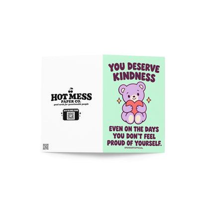 Heartfelt encouragement greeting card featuring a cute lavender teddy bear holding a coral pink heart, surrounded by small golden sparkles on a mint-green background. Bold dark plum retro lettering reads “You Deserve Kindness Even on the Days You Don't Feel Proud of Yourself.” Gentle self-love and self-compassion card by Hot Mess Paper Co.