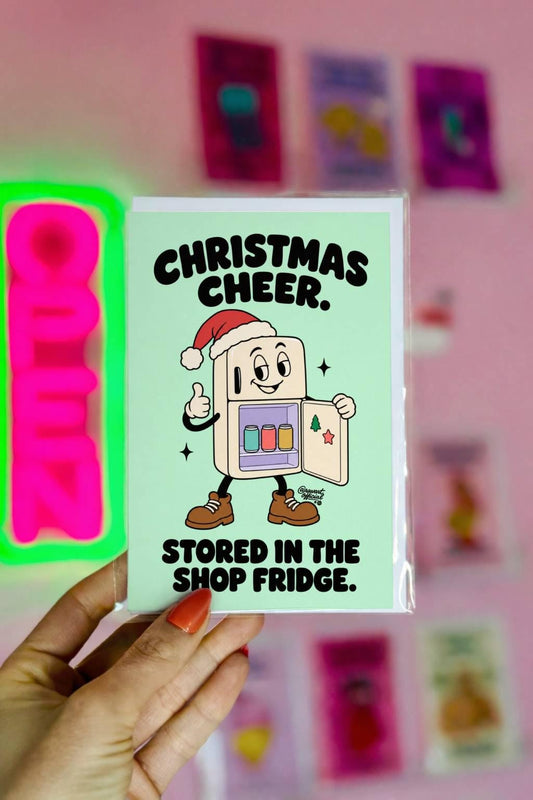 Funny Christmas greeting card featuring a smiling cartoon refrigerator wearing a red Santa hat and brown work boots, giving a thumbs-up while displaying three colourful canned drinks inside its open door. Mint-green background with bold black retro lettering reading “Christmas Cheer. Stored in the Shop Fridge.” Humorous holiday card for mechanics, garage dads, husbands, and workshop enthusiasts.