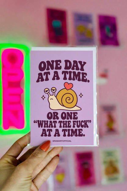 Funny encouragement greeting card featuring a cute cream-coloured cartoon snail with a mustard-yellow spiral shell and a little coral heart on top. Pastel lavender background with dark purple retro lettering reading “One Day at a Time. Or One ‘What the Fuck’ at a Time.” Sarcastic mental health support card with sweary humour.