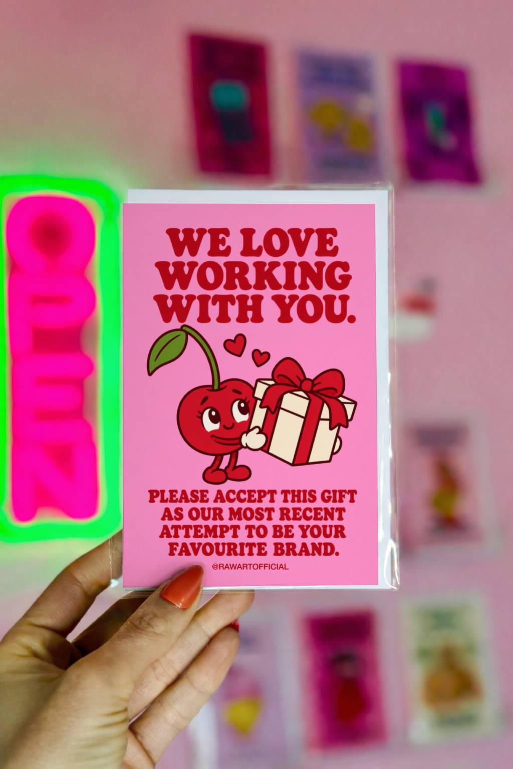 Pink influencer appreciation greeting card with bold red retro lettering reading “We Love Working With You. Please Accept This Gift as Our Most Recent Attempt to Be Your Favourite Brand.” Features a cute smiling red cartoon cherry holding a white gift box tied with a red ribbon, with two floating hearts. A playful brand collaboration thank you card for content creators and influencer PR packages.