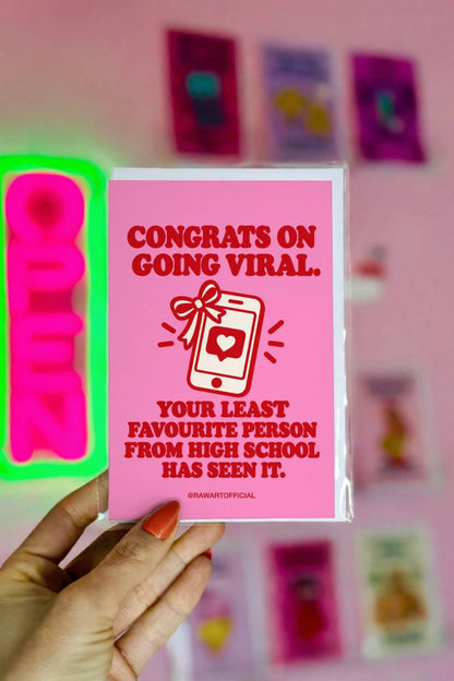 Bright pink funny congratulations greeting card with bold red retro lettering reading “Congrats on Going Viral. Your Least Favourite Person From High School Has Seen It.” Features a white smartphone illustration tied with a red celebration bow, displaying a heart notification icon. A humorous social media milestone card for influencers, TikTok creators, Instagram personalities, and anyone celebrating a viral post.
