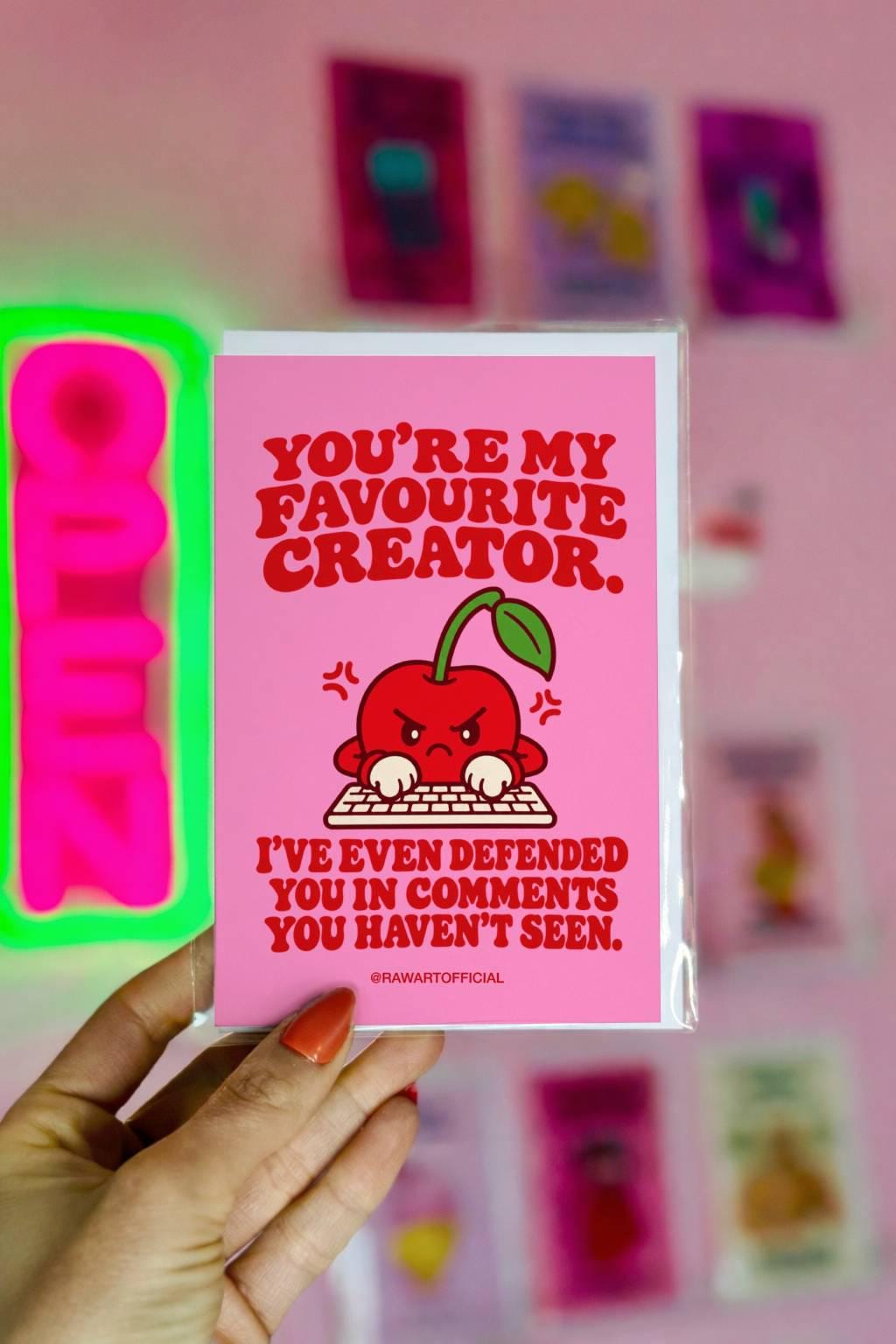 Funny content creator appreciation greeting card featuring an angry red cartoon cherry typing at a computer keyboard on a bright pink background. Bold red retro lettering reads “You're My Favourite Creator. I've Even Defended You in Comments You Haven't Seen.” A humorous influencer thank you card for TikTokers, YouTubers, Instagram creators, and social media personalities.