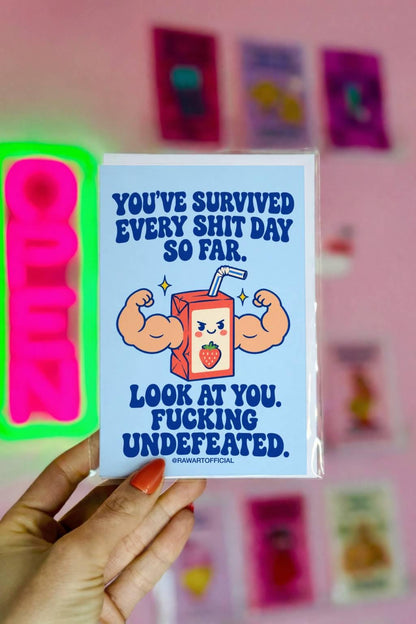 Funny encouragement greeting card featuring a smiling cartoon strawberry juice box with enormous flexing muscular arms, a striped straw, and yellow sparkles. Pastel blue background with bold dark blue retro lettering reading “You've Survived Every Shit Day So Far. Look at You. Fucking Undefeated.” Sarcastic motivational card by Hot Mess Paper Co.