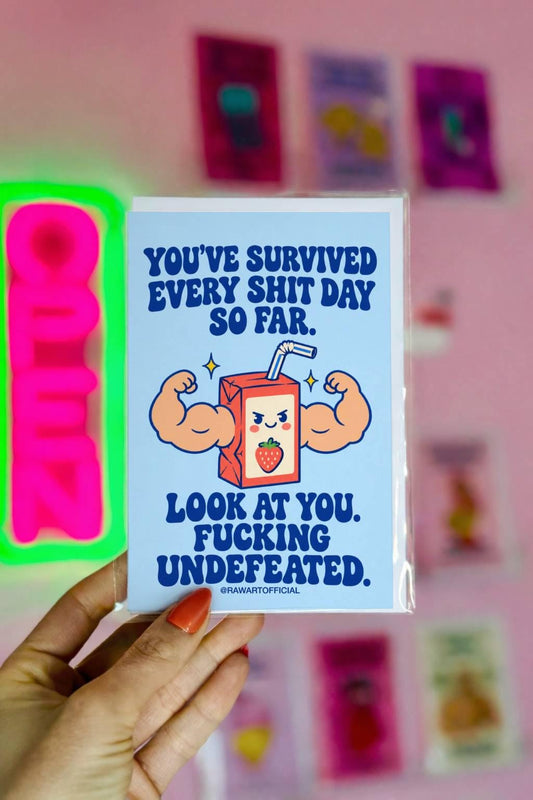 Funny encouragement greeting card featuring a smiling cartoon strawberry juice box with enormous flexing muscular arms, a striped straw, and yellow sparkles. Pastel blue background with bold dark blue retro lettering reading “You've Survived Every Shit Day So Far. Look at You. Fucking Undefeated.” Sarcastic motivational card by Hot Mess Paper Co.