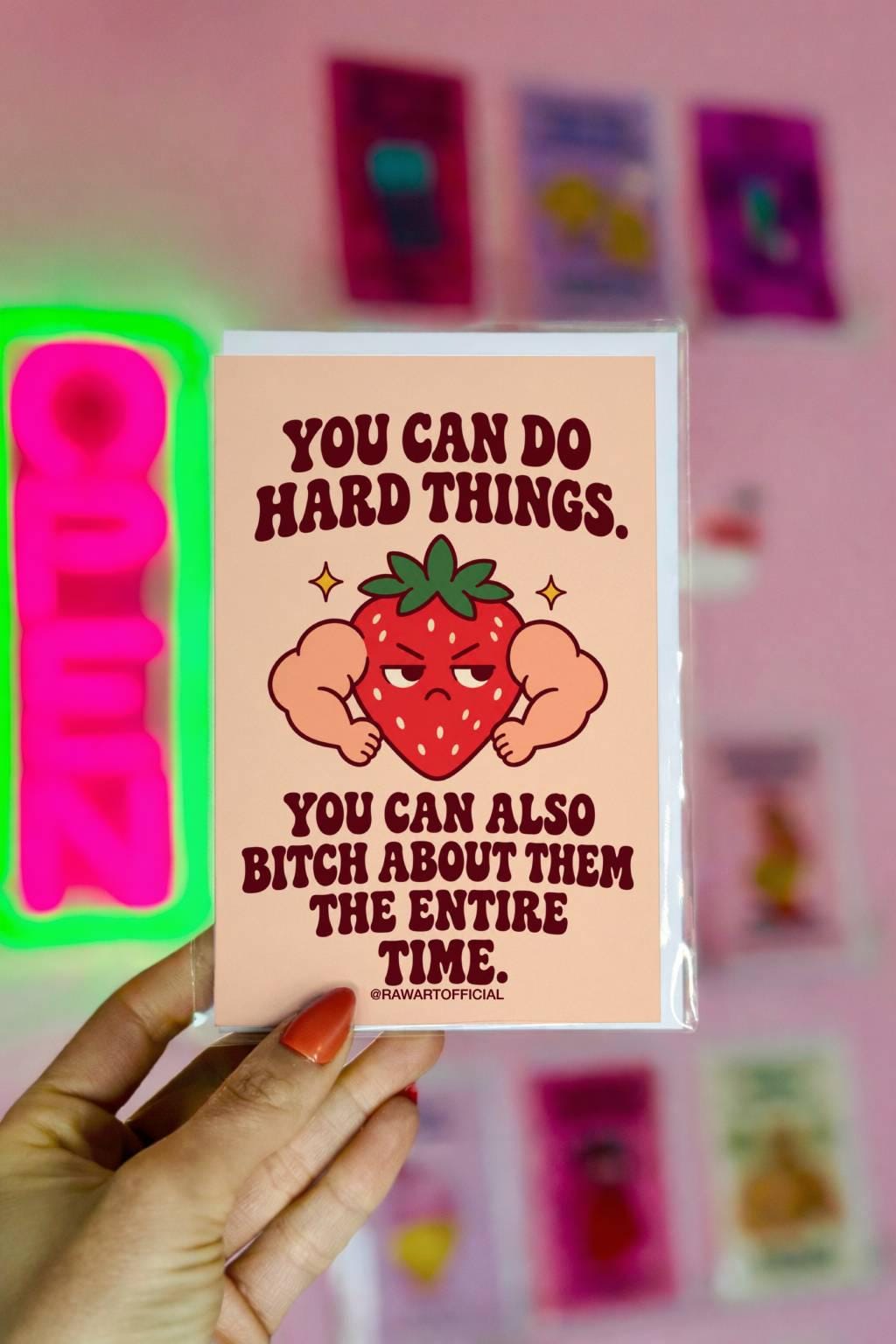 Funny encouragement greeting card featuring a grumpy red cartoon strawberry with oversized flexing muscular arms, green leaves, and golden sparkles. Soft peach background with bold dark burgundy retro lettering reading “You Can Do Hard Things. You Can Also Bitch About Them the Entire Time.” Sarcastic motivational card by Hot Mess Paper Co.