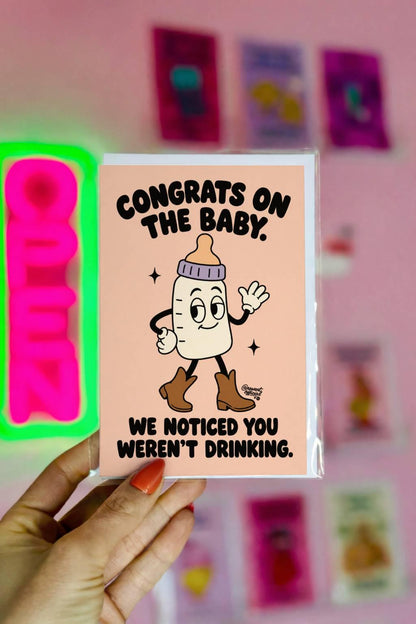 Funny pregnancy congratulations greeting card featuring a smiling cartoon baby bottle with a lavender collar, white gloves, and brown cowboy boots, waving on a peach background. Bold black retro lettering reads “Congrats on the Baby. We Noticed You Weren't Drinking.” Cheeky pregnancy announcement and expecting-parent congratulations card by Hot Mess Paper Co.