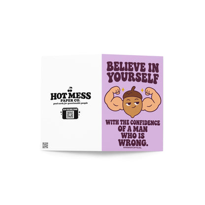 Funny encouragement greeting card with a pastel lavender background and bold dark purple retro lettering reading “Believe in Yourself With the Confidence of a Man Who Is Wrong.” Features a smug cartoon acorn flexing oversized muscular arms with golden sparkles. Sarcastic motivational humor by Hot Mess Paper Co.