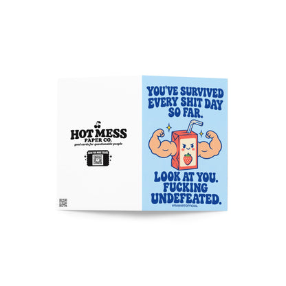 Funny encouragement greeting card featuring a smiling cartoon strawberry juice box with enormous flexing muscular arms, a striped straw, and yellow sparkles. Pastel blue background with bold dark blue retro lettering reading “You've Survived Every Shit Day So Far. Look at You. Fucking Undefeated.” Sarcastic motivational card by Hot Mess Paper Co.