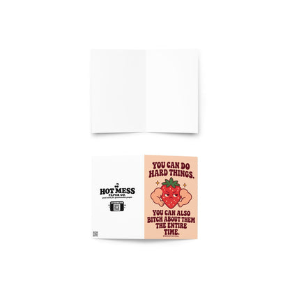 Funny encouragement greeting card featuring a grumpy red cartoon strawberry with oversized flexing muscular arms, green leaves, and golden sparkles. Soft peach background with bold dark burgundy retro lettering reading “You Can Do Hard Things. You Can Also Bitch About Them the Entire Time.” Sarcastic motivational card by Hot Mess Paper Co.