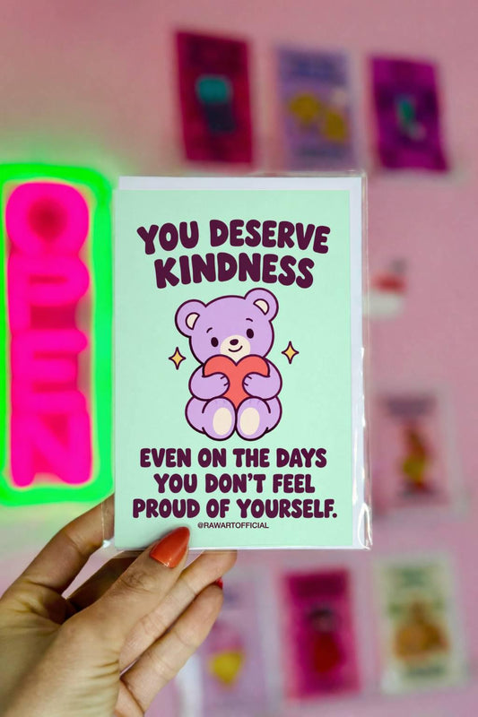 Heartfelt encouragement greeting card featuring a cute lavender teddy bear holding a coral pink heart, surrounded by small golden sparkles on a mint-green background. Bold dark plum retro lettering reads “You Deserve Kindness Even on the Days You Don't Feel Proud of Yourself.” Gentle self-love and self-compassion card by Hot Mess Paper Co.