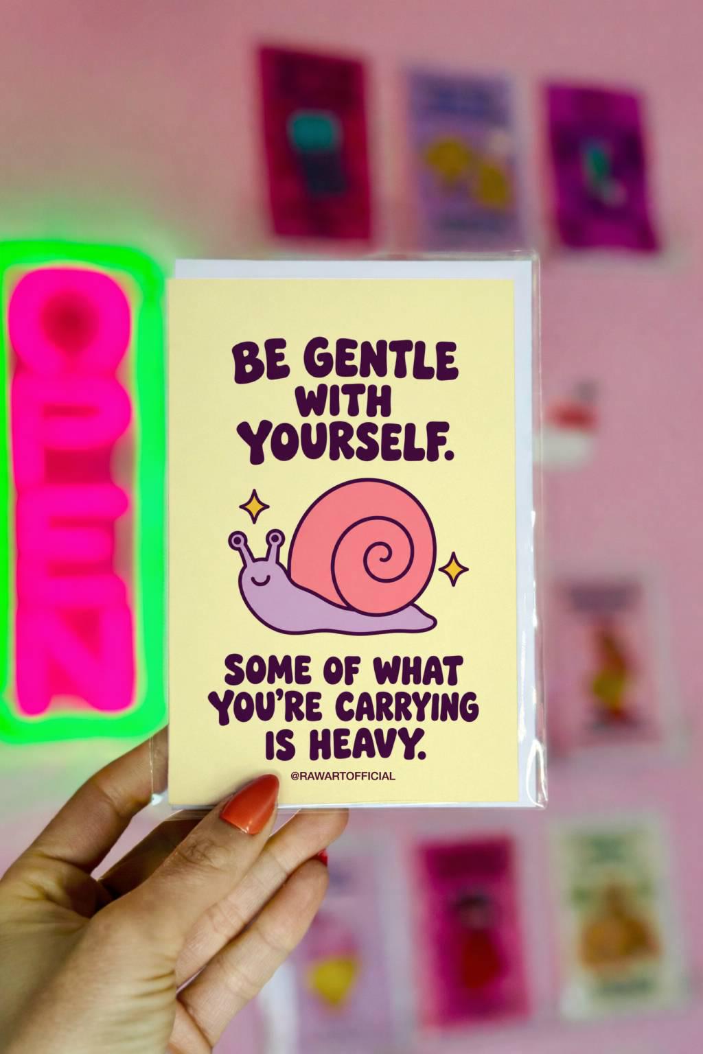 Heartfelt encouragement greeting card with a pastel yellow background and bold dark plum retro lettering reading “Be Gentle With Yourself. Some of What You're Carrying Is Heavy.” Features a smiling lavender snail with a large pink spiral shell and two golden sparkles. Gentle thinking-of-you and emotional support card by Hot Mess Paper Co.