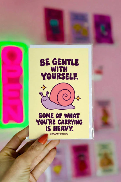 Heartfelt encouragement greeting card with a pastel yellow background and bold dark plum retro lettering reading “Be Gentle With Yourself. Some of What You're Carrying Is Heavy.” Features a smiling lavender snail with a large pink spiral shell and two golden sparkles. Gentle thinking-of-you and emotional support card by Hot Mess Paper Co.