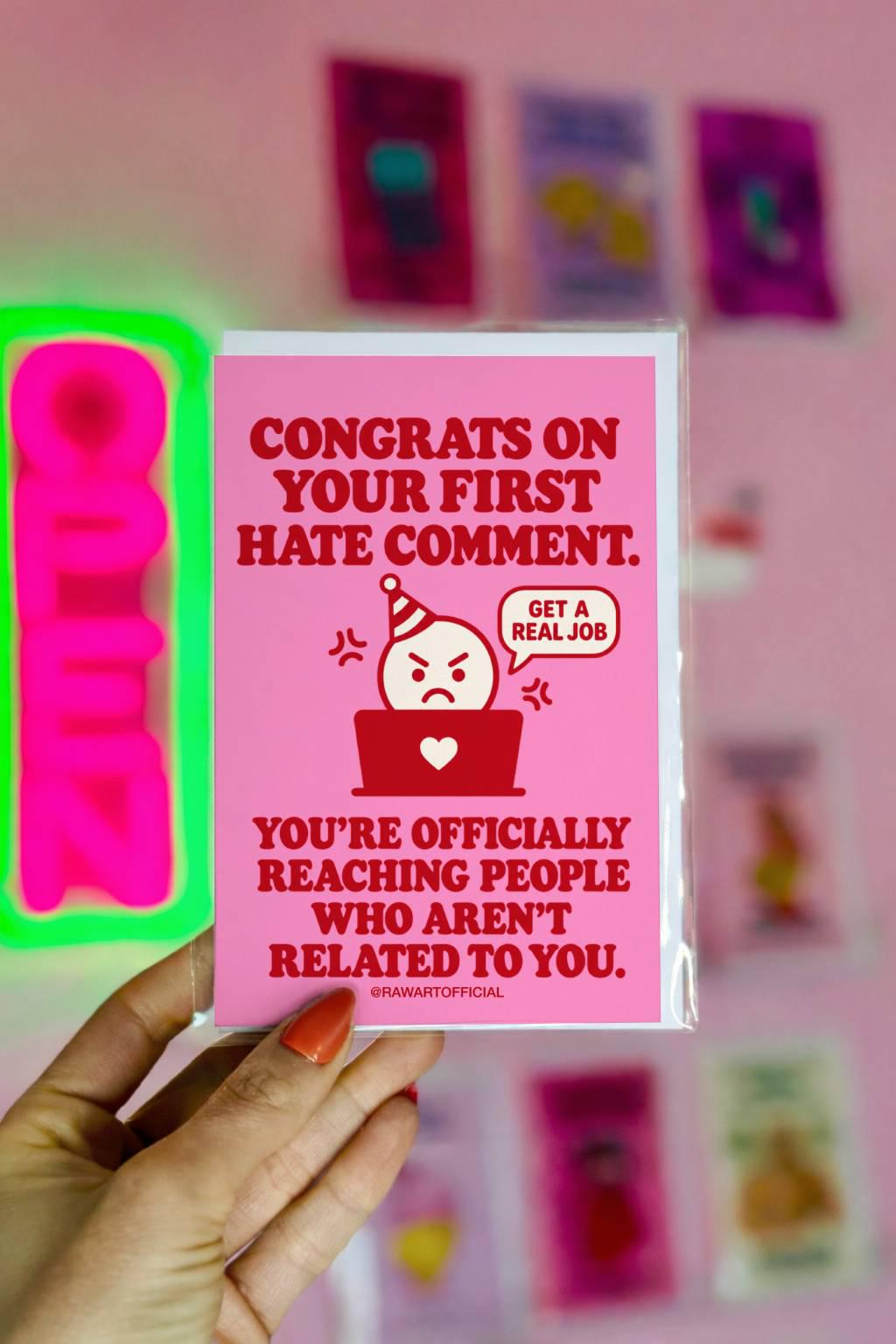 Funny content creator congratulations greeting card with a bright pink background and bold red retro lettering reading “Congrats on Your First Hate Comment. You're Officially Reaching People Who Aren't Related to You.” Features an angry cartoon character wearing a party hat behind a red laptop with a white heart, beside a speech bubble saying “Get a Real Job.” Humorous influencer milestone card by Hot Mess Paper Co.