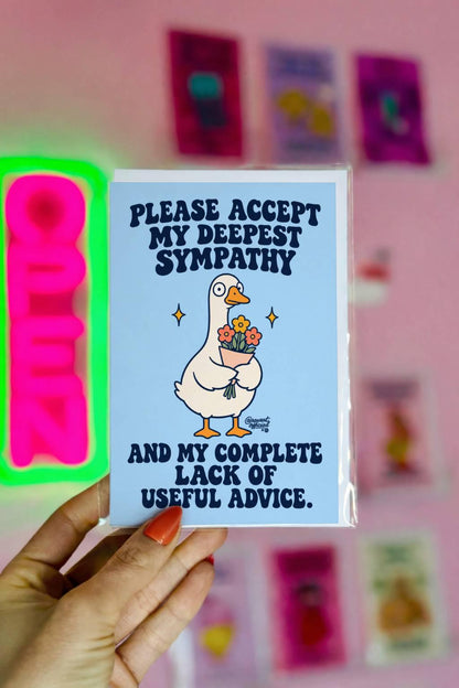 Funny sympathy greeting card featuring a cute white cartoon goose with an orange beak and feet holding a bouquet of pink and orange flowers, surrounded by golden sparkles on a pastel blue background. Bold navy retro lettering reads “Please Accept My Deepest Sympathy and My Complete Lack of Useful Advice.” Unconventional condolence card by Hot Mess Paper Co.