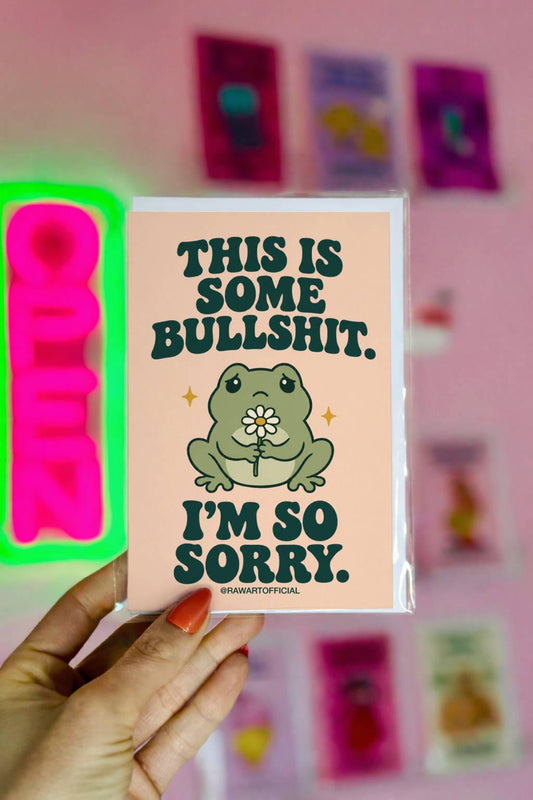 Unconventional sympathy greeting card featuring a sad green cartoon frog holding a small white daisy with a yellow centre. Soft peach background with bold dark green retro lettering reading “This Is Some Bullshit. I'm So Sorry.” A sweary but heartfelt sympathy card for grief, loss, and difficult life situations.