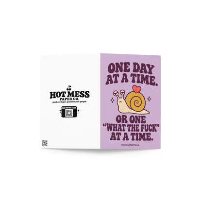 Funny encouragement greeting card featuring a cute cream-coloured cartoon snail with a mustard-yellow spiral shell and a little coral heart on top. Pastel lavender background with dark purple retro lettering reading “One Day at a Time. Or One ‘What the Fuck’ at a Time.” Sarcastic mental health support card with sweary humour.