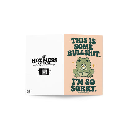 Unconventional sympathy greeting card featuring a sad green cartoon frog holding a small white daisy with a yellow centre. Soft peach background with bold dark green retro lettering reading “This Is Some Bullshit. I'm So Sorry.” A sweary but heartfelt sympathy card for grief, loss, and difficult life situations.