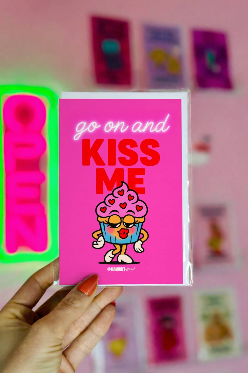 Funny greeting card reading “Go On and Kiss Me” with a cartoon cupcake blowing a kiss on a bright pink background.