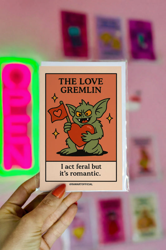 Green gremlin holding a big red heart and love flag with the phrase “I act feral but it’s romantic.”