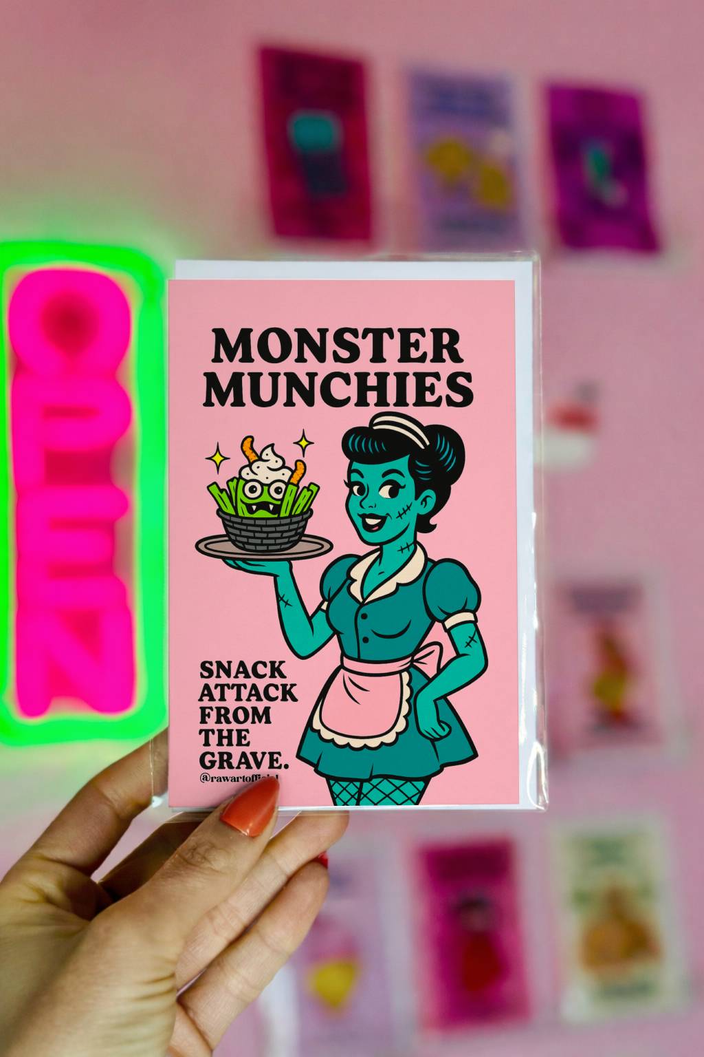 Greeting card with a pink background and a cartoon illustration. The illustration features a green-skinned waitress holding a tray with a basket of monster snacks. Text reads "Monster Munchies" and "Snack Attack From The Grave."