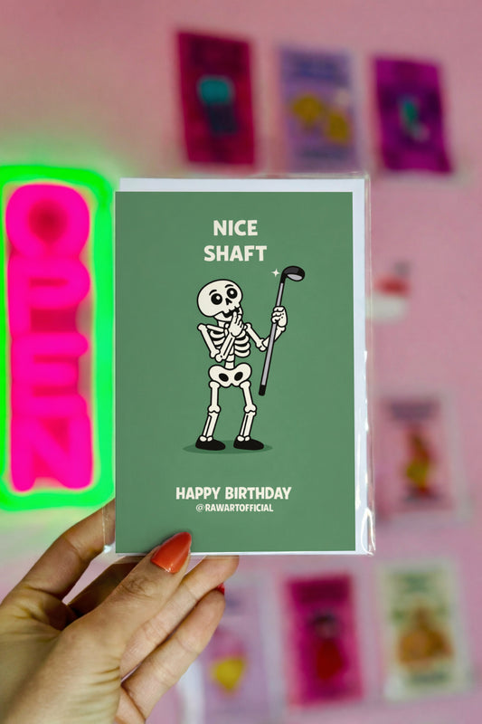Hand holding a birthday card with a skeleton holding a golf club and text 'Nice Shaft' on a green background.
