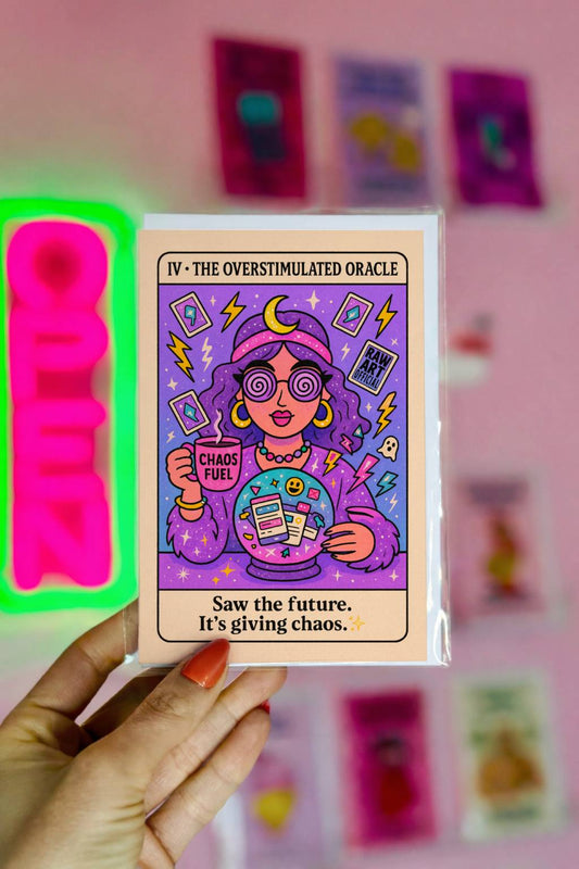 Tarot-style greeting card featuring a purple-haired oracle with spiral glasses holding a crystal ball filled with phone notifications and a mug labeled “Chaos Fuel,” surrounded by lightning bolts and stars.