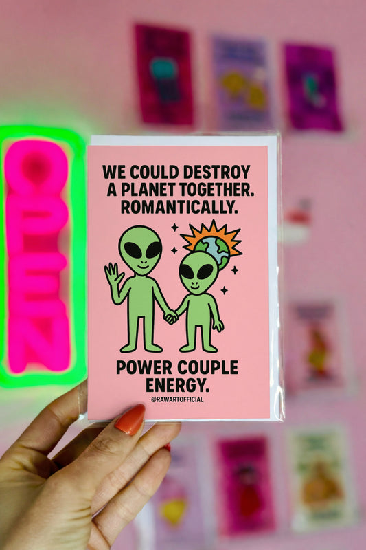 Alien couple holding hands with a tiny exploding planet behind them on a pink background. Text says “We could destroy a planet together. Romantically.” Funny Valentine card.
