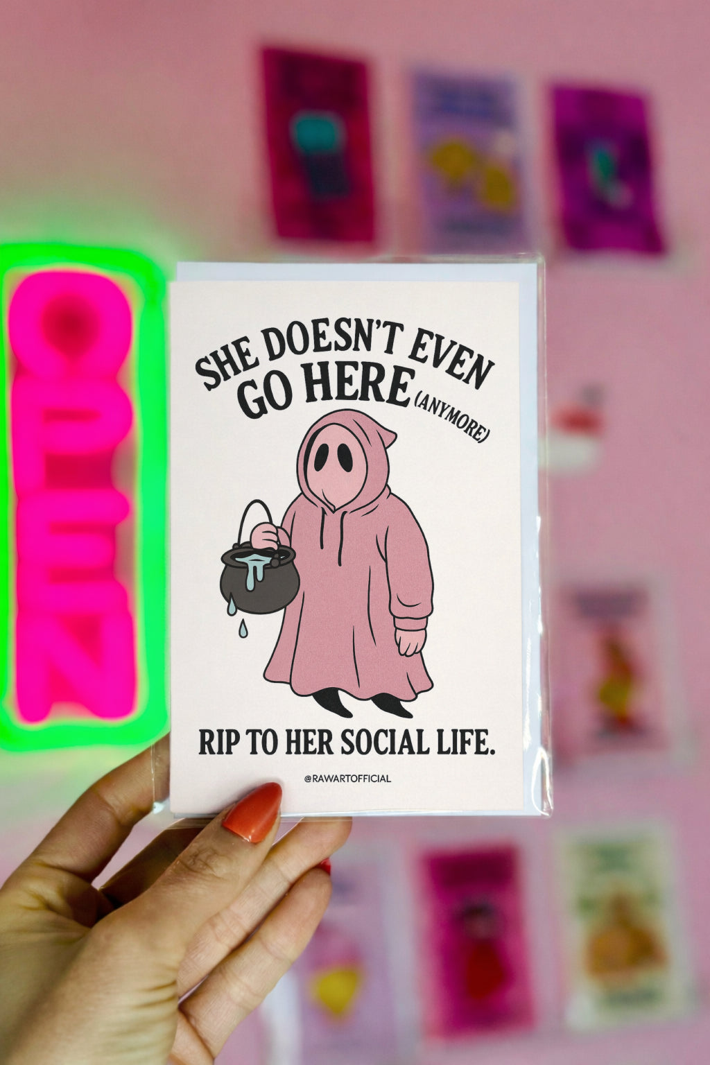 Pink ghost in a hoodie holding a cauldron with text “She Doesn’t Even Go Here (Anymore)” – funny Mean Girls inspired Halloween card.