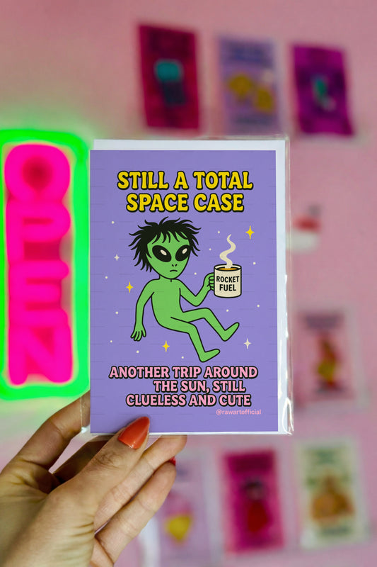Green alien holding a coffee mug labeled “Rocket Fuel” floating in space with text “Still a Total Space Case.”