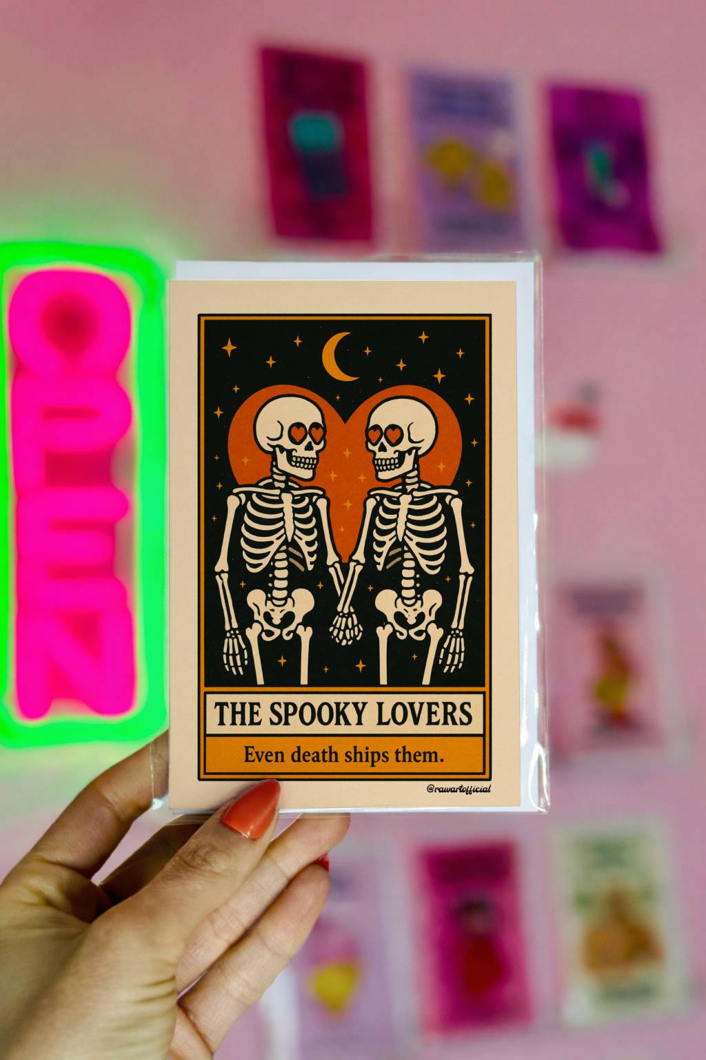 Tarot-style greeting card featuring two skeletons with heart-shaped eyes holding hands under a crescent moon, framed by an orange heart on a black starry background with the text “The Spooky Lovers – Even death ships them.”