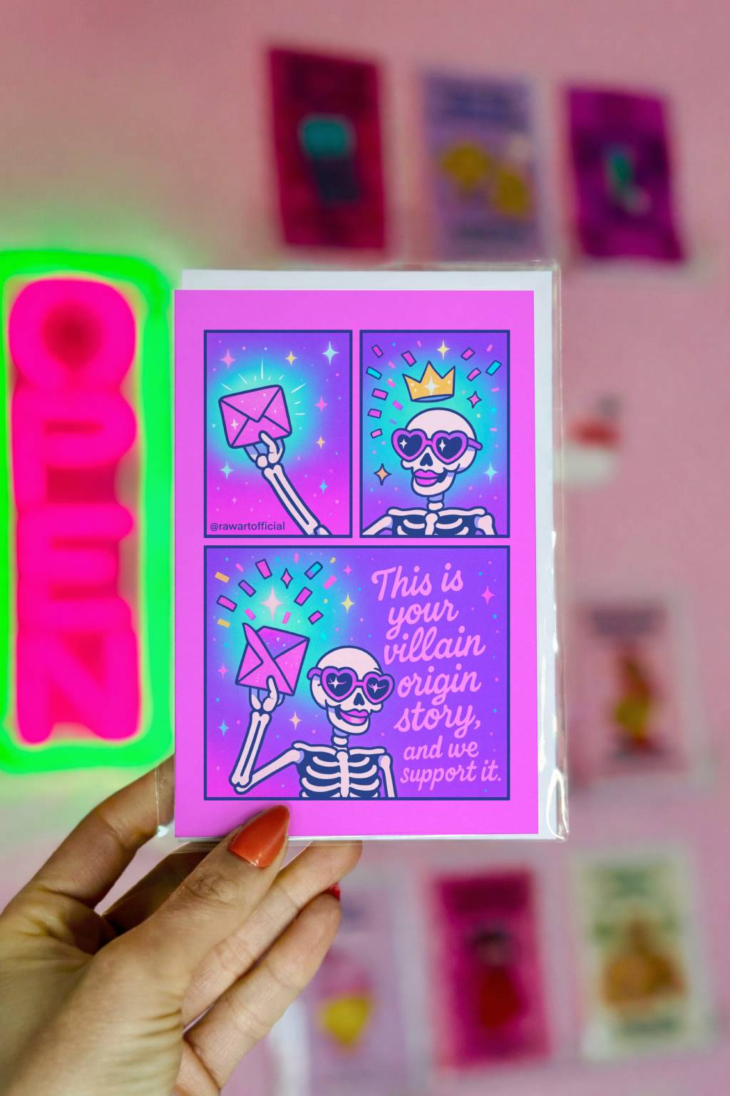 Retro comic-style greeting card featuring a glam skeleton holding a pink envelope with the text “This is your villain origin story, and we support it.”