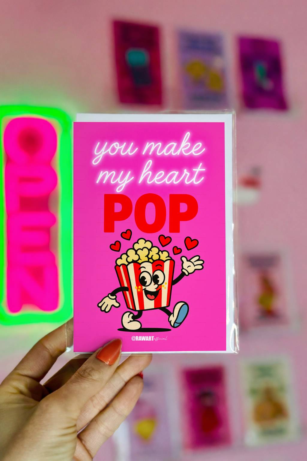 Bright pink greeting card reading You Make My Heart Pop with smiling popcorn illustration and @rawartofficial signature.